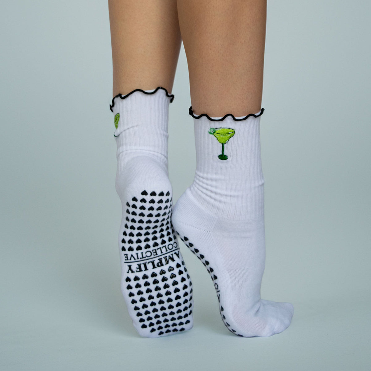 White ankle socks worn on a model's feet, featuring a green margarita glass graphic on the cuff, black ruffled trim, and black grip dots on the sole. The brand name 'AMPLIFY COLLECTIVE' is printed on the sole.