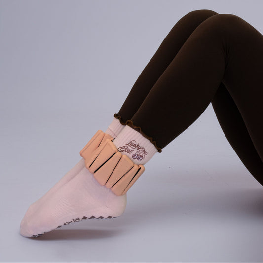 A close-up of a person's legs and foot wearing a pale pink sock with a wide beige ankle cuff and dark brown leggings, set against a solid light gray studio background.