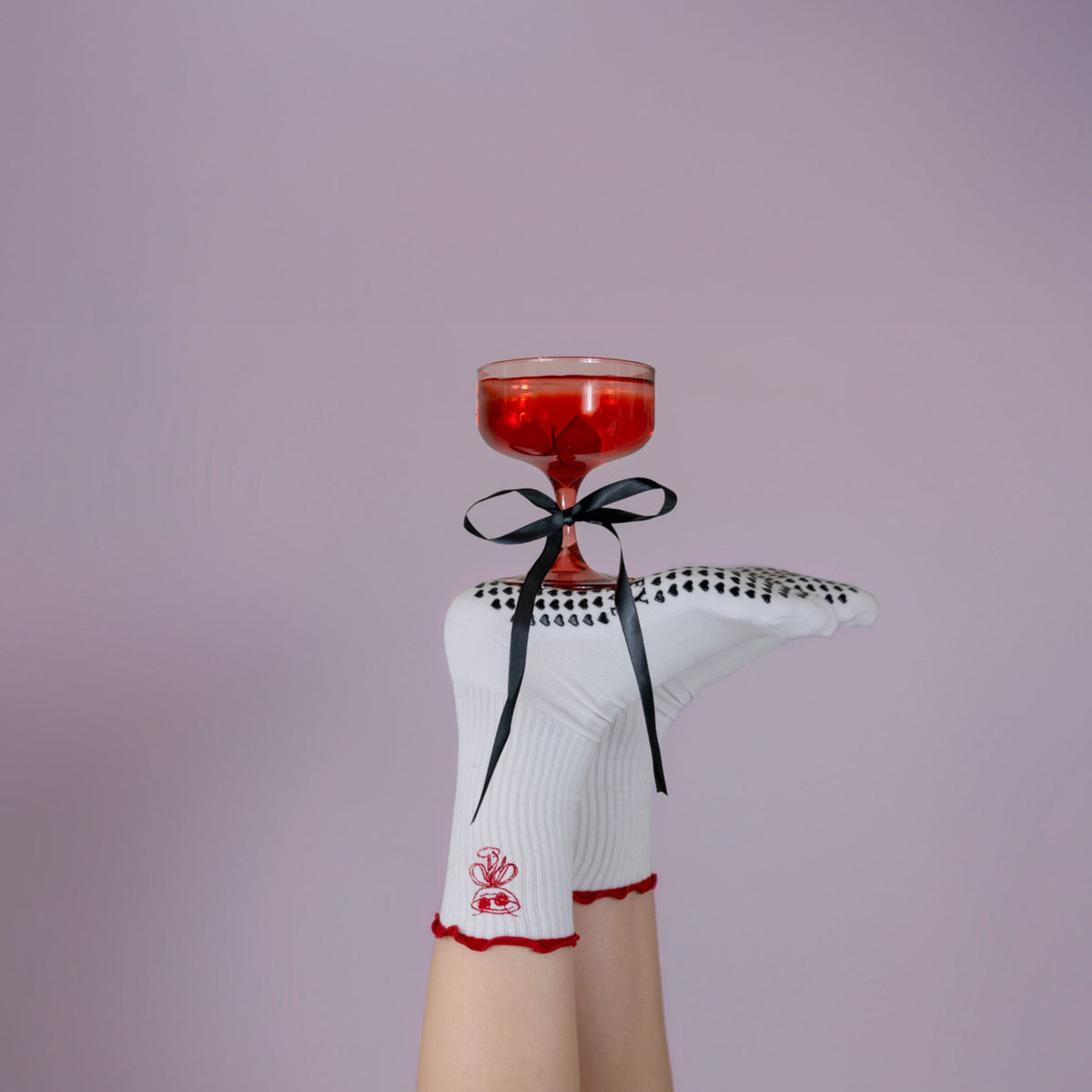 Cocktail glass with red liquid held by a person wearing a white outfit with black and red details on a purple background
